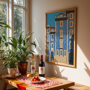 K&ouml;nnte beinhalten: Ein gerahmter Druck eines blauen Geb&auml;udes mit wei&szlig;en und gelben Akzenten h&auml;ngt an einer Wand. Auf einem Holztisch stehen eine Wei&szlig;weinflasche, eine Rotweinflasche und eine Sch&uuml;ssel mit Tomaten. Topfpflanzen stehen in der N&auml;he eines Fensters.