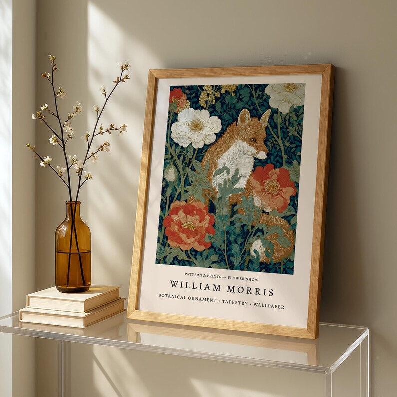 a framed print of a fox surrounded by flowers, placed on a glass table next to a vase with white flowers.