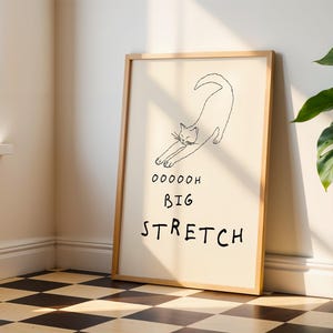 May include: A framed art print featuring a minimalist line drawing of a stretching cat. The artwork includes the words "Ooooooh Big Stretch" in black font. The frame is a light wood color, and the background is a cream color.