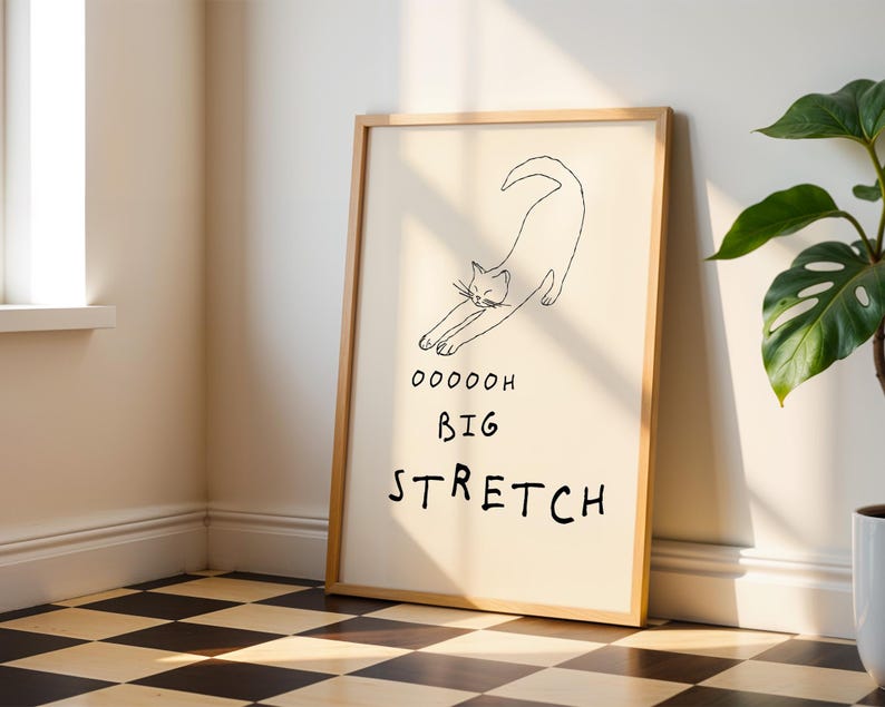 May include: A framed art print featuring a minimalist line drawing of a stretching cat. The artwork includes the words "Ooooooh Big Stretch" in black font. The frame is a light wood color, and the background is a cream color.