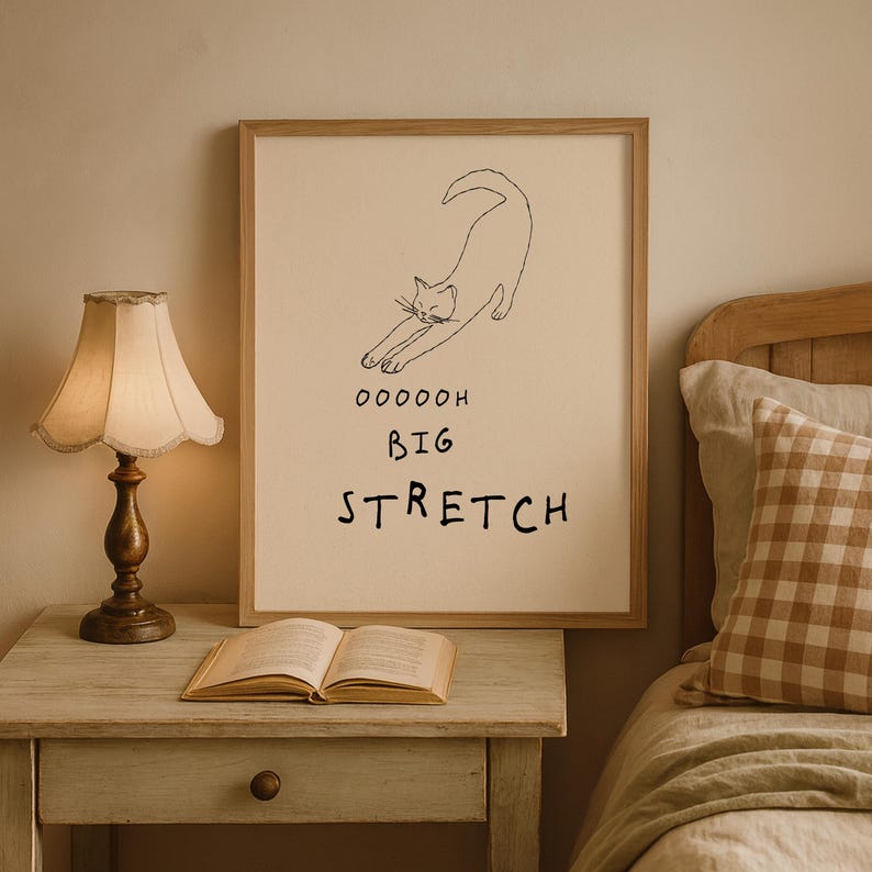 May include: A framed print featuring a minimalist line drawing of a stretching cat. The text "OOOOOH BIG STRETCH" is below the cat illustration. The artwork is displayed in a bedroom setting, with a lamp and an open book on a nightstand.