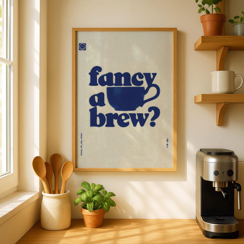May include: A framed poster with the text "fancy a brew?" in dark blue, featuring a coffee cup graphic. The poster is in a light-colored wooden frame, displayed in a kitchen setting with wooden spoons and a coffee maker.