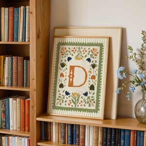 May include: A framed art print featuring a large letter "D" in a warm orange hue, surrounded by a floral design in green, orange, and blue. The artwork is displayed on a wooden shelf with a backdrop of a bookshelf filled with books.