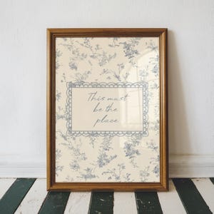Puede incluir: Un estampado enmarcado con un diseño floral azul y crema. El estampado presenta el texto "This must be the place" dentro de un borde decorativo. El marco es de madera.