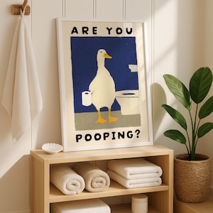 May include: A framed art print featuring a white duck holding toilet paper in a bathroom setting. The text on the print reads "ARE YOU POOPING?" in black capital letters. The print is displayed above a wooden shelf with rolled towels.