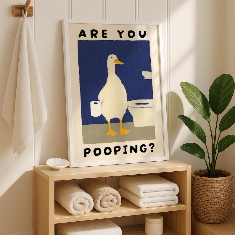 May include: A framed art print featuring a white duck holding toilet paper in a bathroom setting. The text on the print reads "ARE YOU POOPING?" in black capital letters. The print is displayed above a wooden shelf with rolled towels.