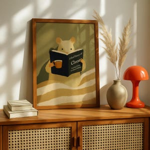 May include: A framed print shows a cartoon mouse reading "A Brief History of Cheese" and holding a mug. The artwork has a warm colour palette and a wooden frame. A small stack of books, a vase with dried plants, and an orange lamp are on a wooden cabinet.