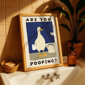 May include: A framed art print featuring a cartoon duck in a bathroom setting. The duck is white with yellow feet and a yellow beak, holding toilet paper. The text "ARE YOU POOPING?" is displayed above and below the duck illustration.