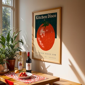 a framed poster of a tomato with the text "Kitchen Disco" and "A little sauce for good food and bad dancing" on it, along with a wooden table with a bowl of fruit, a bottle of wine, and a potted plant.
