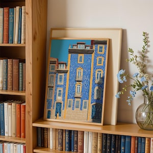 K&ouml;nnte beinhalten: Ein gerahmter Kunstdruck eines blauen Geb&auml;udes mit gelben Details und wei&szlig;en Fenstern. Das Kunstwerk wird auf einem Holzregal neben einer Vase mit blauen Blumen und einem B&uuml;cherregal mit B&uuml;chern ausgestellt.