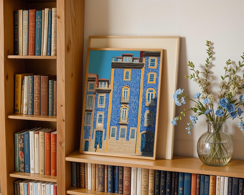 K&ouml;nnte beinhalten: Ein gerahmter Kunstdruck eines blauen Geb&auml;udes mit gelben Details und wei&szlig;en Fenstern. Das Kunstwerk wird auf einem Holzregal neben einer Vase mit blauen Blumen und einem B&uuml;cherregal mit B&uuml;chern ausgestellt.