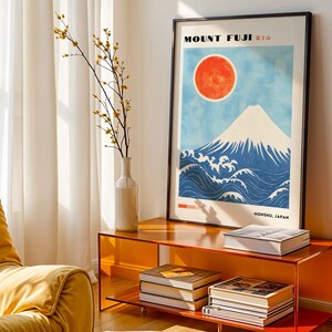 Black-framed Mount Fuji artwork on an orange side table, styled with books and a white vase. Japanese wall art features snow-topped mountain, red sun, and traditional wave design. Perfect mountain print for living room decor.