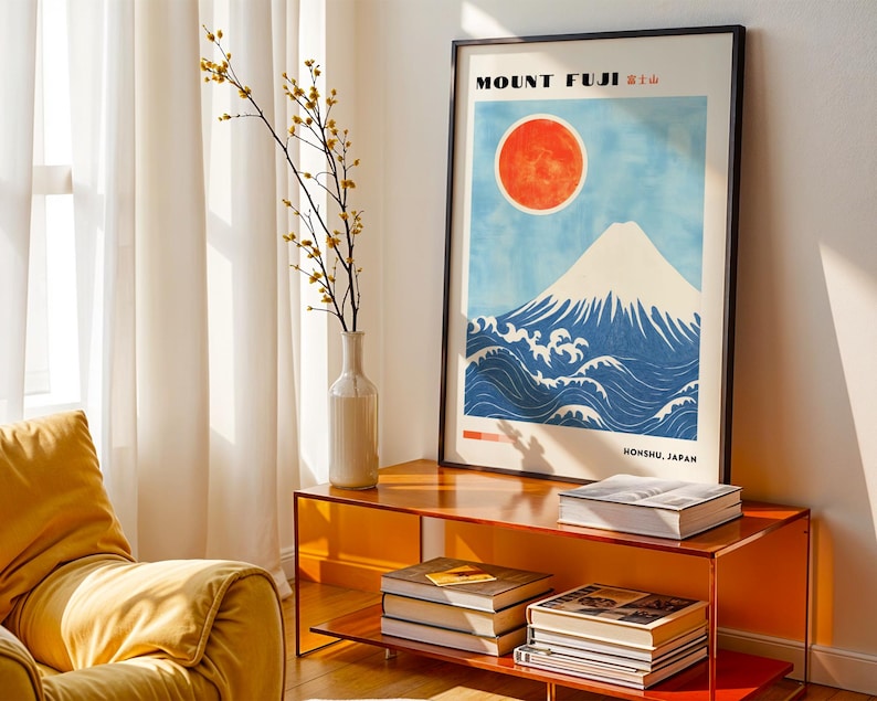 Black-framed Mount Fuji artwork on an orange side table, styled with books and a white vase. Japanese wall art features snow-topped mountain, red sun, and traditional wave design. Perfect mountain print for living room decor.