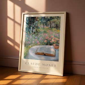 Puede incluir: Impresión artística enmarcada de "Jardin du Matin" de Claude Monet. La obra representa una escena de jardín con flores coloridas, árboles y una mesa con un libro abierto y una pequeña maceta. La impresión está en un marco claro.