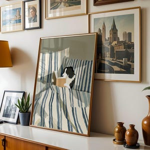 Op de afbeelding: Een ingelijste prent van een hond die in bed leest, met blauw-wit gestreept beddengoed. De prent wordt tentoongesteld met andere ingelijste kunstwerken, een lamp en vazen. De hond is zwart en wit.