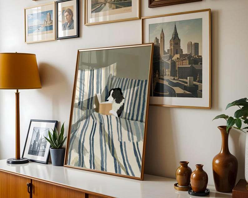 Op de afbeelding: Een ingelijste prent van een hond die in bed leest, met blauw-wit gestreept beddengoed. De prent wordt tentoongesteld met andere ingelijste kunstwerken, een lamp en vazen. De hond is zwart en wit.