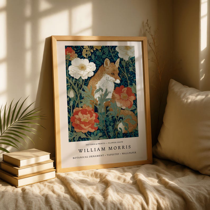 A framed print of a fox surrounded by flowers is displayed on a bed, with a stack of books and a plant nearby.