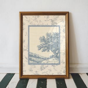 May include: A framed print with a blue and white landscape scene, including a tree. The print is set against a cream background with a floral pattern, enclosed in a brown wooden frame. The style is vintage and evokes a pastoral feel.