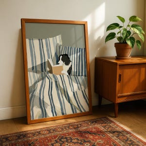 Op de afbeelding: Een ingelijste kunstdruk toont een zwart-witte hond die een boek leest op een bed met blauw-wit gestreepte beddengoed. Het kunstwerk zit in een houten lijst. Een plant in een pot staat op een houten kast.