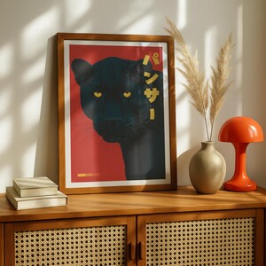May include: Framed art print featuring a black panther on a red background. The panther's yellow eyes stand out against its dark fur. Japanese text is displayed vertically on the right side. The print is in a wooden frame.