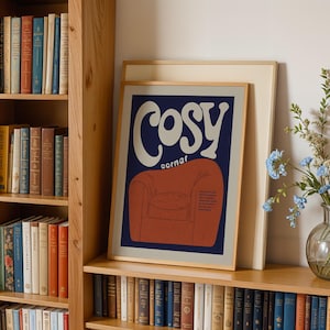 May include: A framed art print with the words "Cosy Corner" in a retro font, featuring an illustration of a red armchair on a navy blue background. The print is displayed on a wooden shelf next to a bookshelf filled with books.