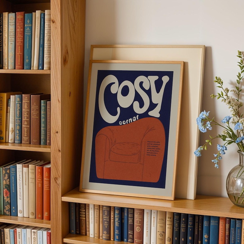 May include: A framed art print with the words "Cosy Corner" in a retro font, featuring an illustration of a red armchair on a navy blue background. The print is displayed on a wooden shelf next to a bookshelf filled with books.
