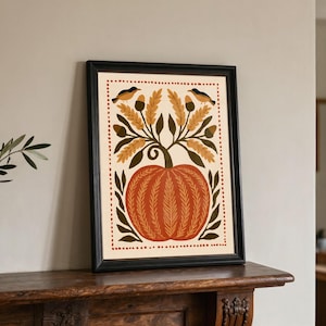 May include: A framed wall art print featuring a central orange pumpkin with textured lines, surrounded by green leaves, golden wheat stalks, and two small birds perched at the top. The design is enclosed within a dotted red rectangular border.