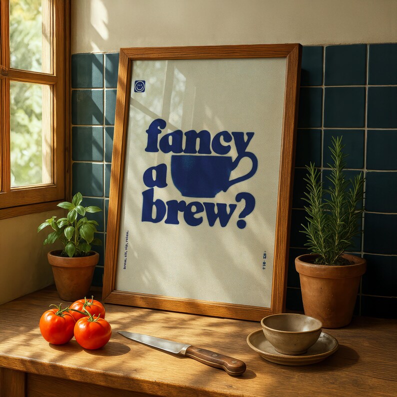 May include: A framed print with the text "fancy a brew?" in blue, featuring a blue coffee cup graphic. The print is in a wooden frame, resting on a wooden surface with tomatoes, a knife, and a small bowl. A potted plant is also visible.