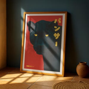 May include: Framed art print featuring a black panther illustration. The panther's face is set against a red background with Japanese text in gold. The print is in a wooden frame, leaning against a dark blue wall.