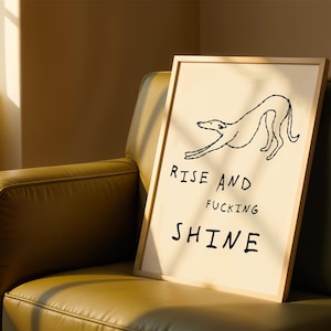 Op de afbeelding: Een ingelijste kunstprint met een minimalistische lijntekening van een hond die zich uitrekt. De tekst "RISE AND FUCKING SHINE" staat in zwarte letters. De print heeft een cr&egrave;me achtergrond en een lichte houten lijst.