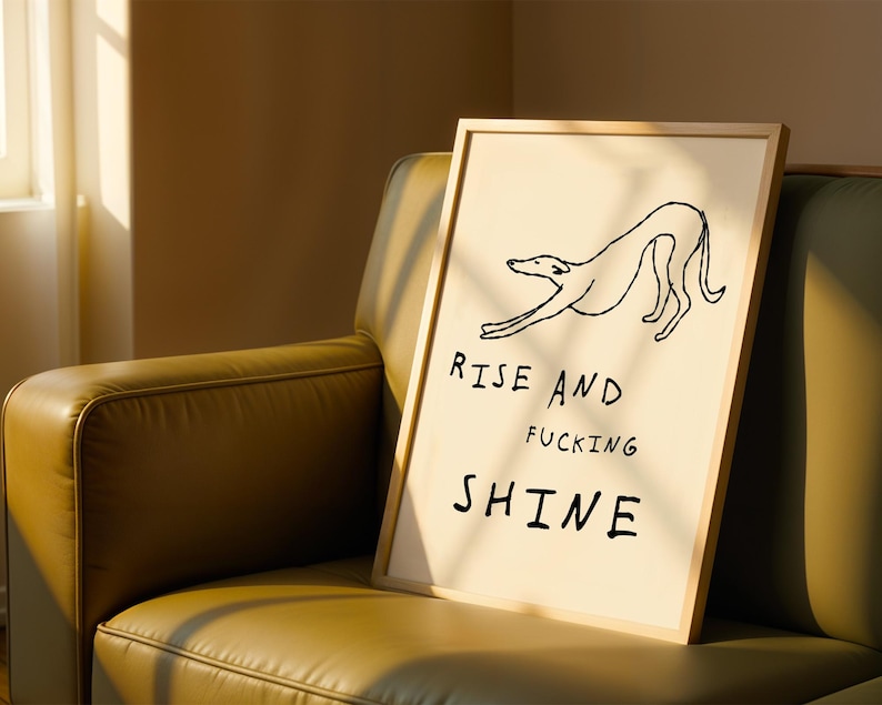 Op de afbeelding: Een ingelijste kunstprint met een minimalistische lijntekening van een hond die zich uitrekt. De tekst "RISE AND FUCKING SHINE" staat in zwarte letters. De print heeft een cr&egrave;me achtergrond en een lichte houten lijst.