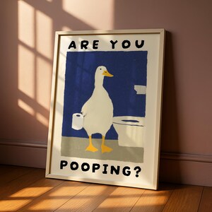 May include: Framed art print featuring a white duck holding toilet paper, standing in front of a toilet. The background is a blue rectangle with the words "ARE YOU POOPING?" in black. The frame is a light beige color.