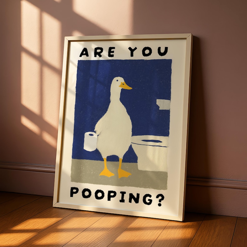 May include: Framed art print featuring a white duck holding toilet paper, standing in front of a toilet. The background is a blue rectangle with the words "ARE YOU POOPING?" in black. The frame is a light beige color.