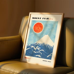 Framed Mount Fuji poster resting on a leather sofa, bathed in warm sunlight. Japanese print features bold blue waves, white peak, and bright red sun. Ideal Japanese-inspired wall art for modern home styling.