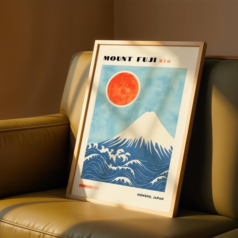 Framed Mount Fuji poster resting on a leather sofa, bathed in warm sunlight. Japanese print features bold blue waves, white peak, and bright red sun. Ideal Japanese-inspired wall art for modern home styling.