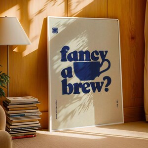May include: A framed poster with the words "fancy a brew?" in blue, along with a blue coffee cup graphic. The poster has a beige background and a white frame. The text "brew, sip, relax." is printed at the bottom.