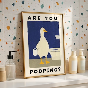 May include: A framed art print featuring a cartoon duck holding toilet paper in front of a toilet. The text "ARE YOU POOPING?" is displayed in black. The artwork has a gold frame and is set against a colorful tile background.