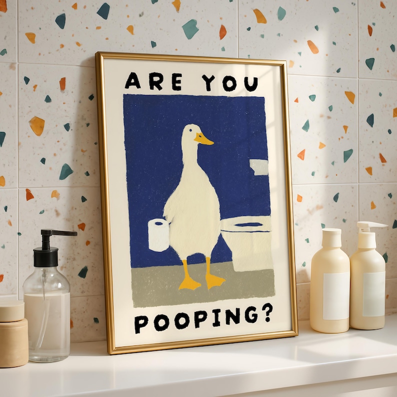 May include: A framed art print featuring a cartoon duck holding toilet paper in front of a toilet. The text "ARE YOU POOPING?" is displayed in black. The artwork has a gold frame and is set against a colorful tile background.