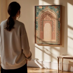 May include: A framed artwork depicting an ornate archway with floral and geometric patterns in shades of pink, green, and gold. The artwork is displayed on a white wall, and a person is standing in front of it, looking at the artwork.