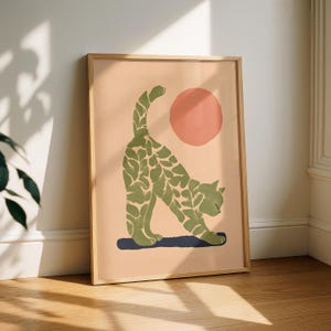 Könnte beinhalten: Gerahmter Kunstdruck mit einer stilisierten, sich streckenden Katze. Die Katze ist grün mit einem blattartigen Muster, vor einem pfirsichfarbenen Hintergrund mit einer korallenfarbenen Sonne. Der Rahmen ist aus Naturholz.