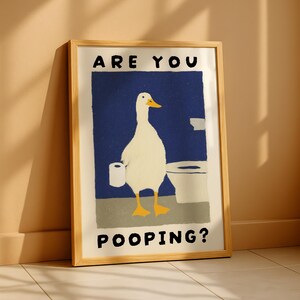 May include: Framed art print featuring a whimsical illustration of a white duck holding toilet paper in a bathroom setting. The artwork has a blue background and the text "ARE YOU POOPING?" in bold, black letters.