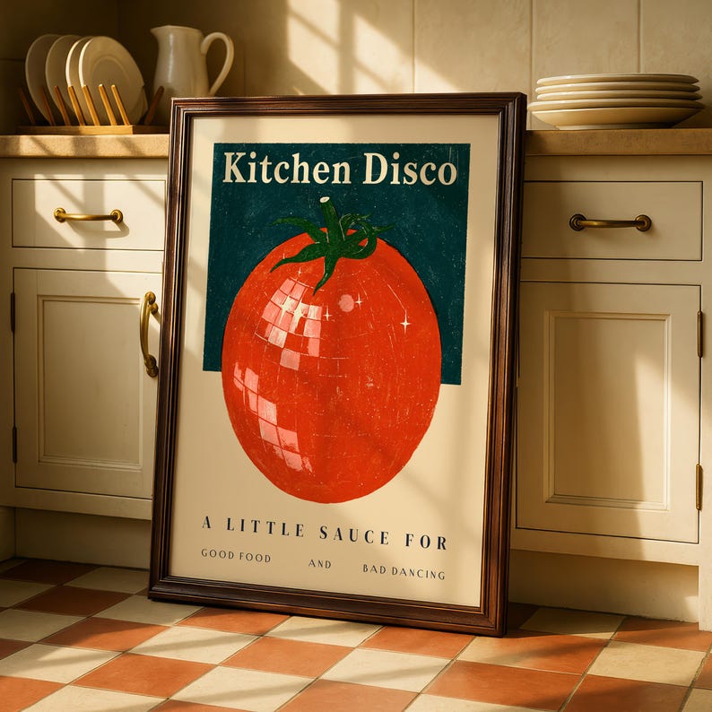 A framed poster with a large tomato graphic and text, placed on a kitchen counter with dishes and cabinets in the background.