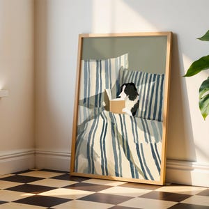 Op de afbeelding: Een ingelijste kunstdruk toont een zwart-witte hond die in bed leest. Het beddengoed heeft blauw-witte strepen. Het kunstwerk staat in een licht houten lijst en leunt tegen een muur. Een plant in een pot staat op de achtergrond.