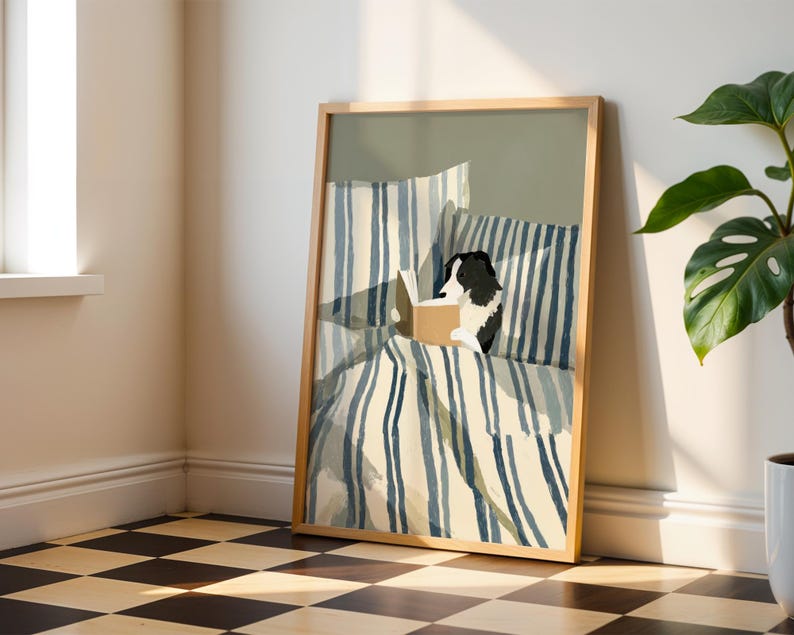 Op de afbeelding: Een ingelijste kunstdruk toont een zwart-witte hond die in bed leest. Het beddengoed heeft blauw-witte strepen. Het kunstwerk staat in een licht houten lijst en leunt tegen een muur. Een plant in een pot staat op de achtergrond.