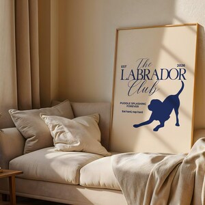 Puede incluir: Un p&oacute;ster enmarcado con fondo crema y texto y gr&aacute;ficos azul marino. El texto dice "The Labrador Club" con una silueta de un perro labrador. El p&oacute;ster se muestra en un sof&aacute; beige.