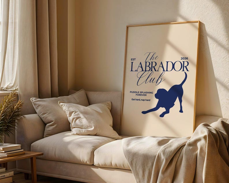 Puede incluir: Un p&oacute;ster enmarcado con fondo crema y texto y gr&aacute;ficos azul marino. El texto dice "The Labrador Club" con una silueta de un perro labrador. El p&oacute;ster se muestra en un sof&aacute; beige.