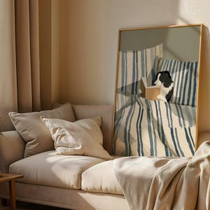 Op de afbeelding: Een ingelijste kunstdruk toont een zwart-witte hond die in bed leest. Het kunstwerk toont blauw-wit gestreepte beddengoed en kussens. De print wordt weergegeven boven een beige bank met bijpassende kussens en een houten bijzettafel.