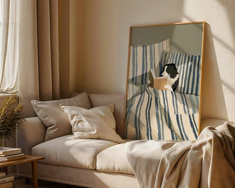 Op de afbeelding: Een ingelijste kunstdruk toont een zwart-witte hond die in bed leest. Het kunstwerk toont blauw-wit gestreepte beddengoed en kussens. De print wordt weergegeven boven een beige bank met bijpassende kussens en een houten bijzettafel.