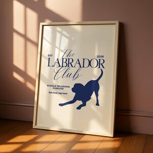 Puede incluir: Un estampado enmarcado con fondo crema y marco dorado. El estampado presenta el texto "The Labrador Club" en escritura azul marino, junto con una silueta azul marino de un perro Labrador. Tambi&eacute;n se incluye el texto "Puddle splashing forever" y "Eat hard, nap hard".
