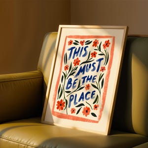 Puede incluir: Una impresión artística enmarcada con las palabras "THIS MUST BE THE PLACE" en azul. La obra presenta flores rojas y hojas verdes sobre un fondo crema, enmarcada por un marco de madera clara.